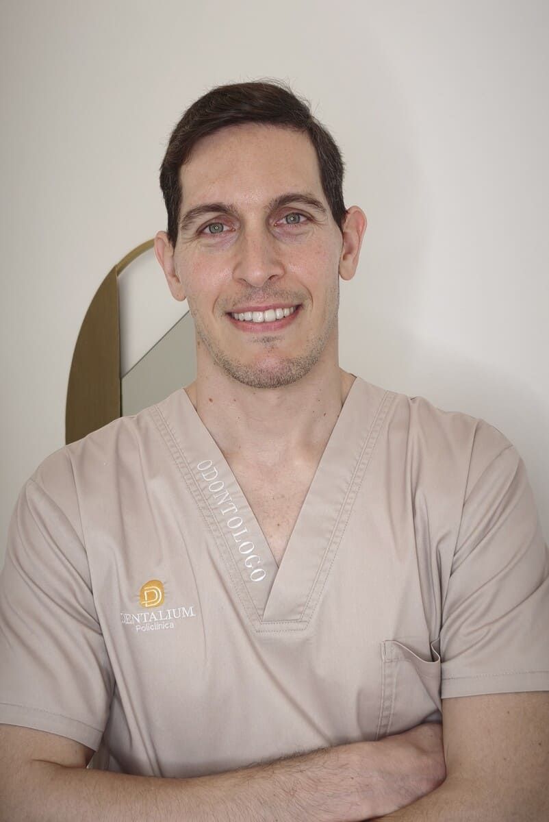 Hombre con uniforme médico beige sonriendo con los brazos cruzados contra una pared lisa.