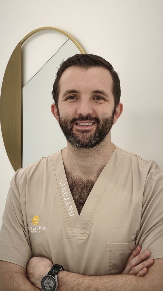 Hombre con uniforme quirúrgico beige sonriendo con los brazos cruzados en un entorno clínico.