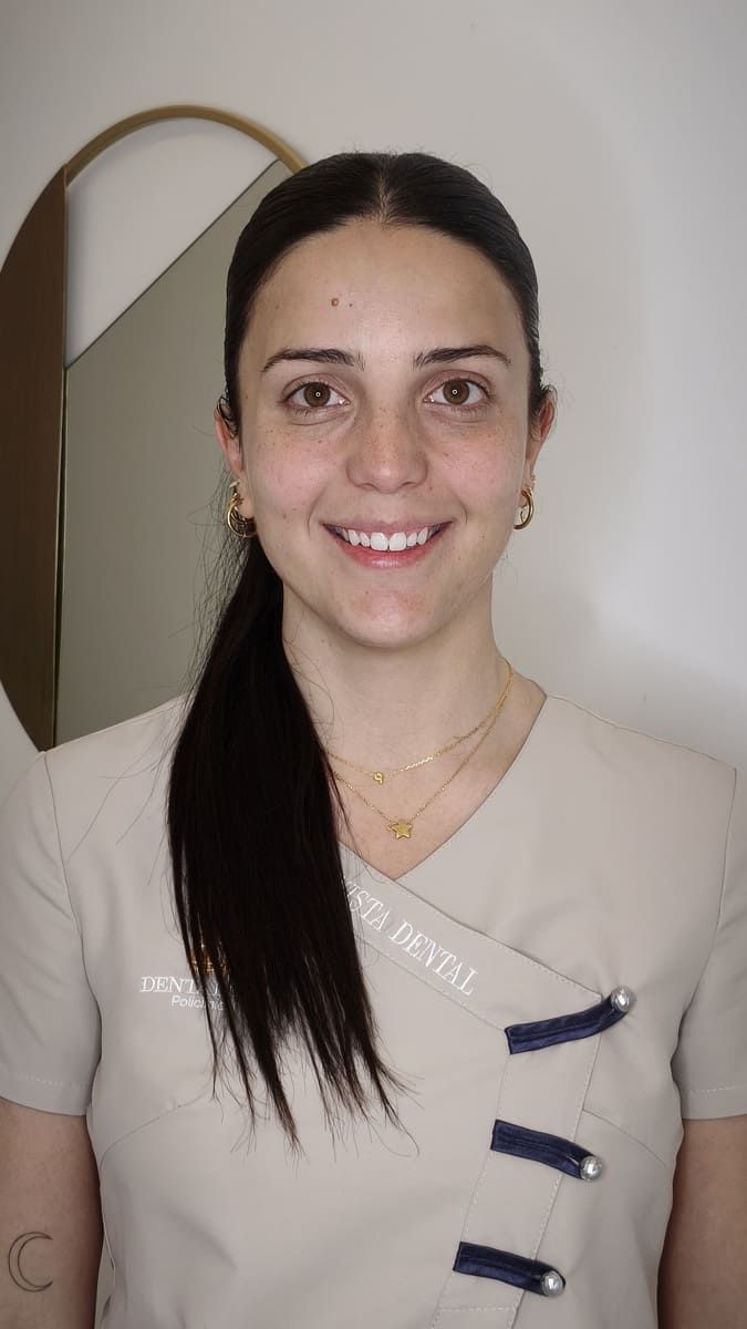 Mujer sonriente con uniforme beige de pie en el interior de una casa, con una pared clara de fondo.