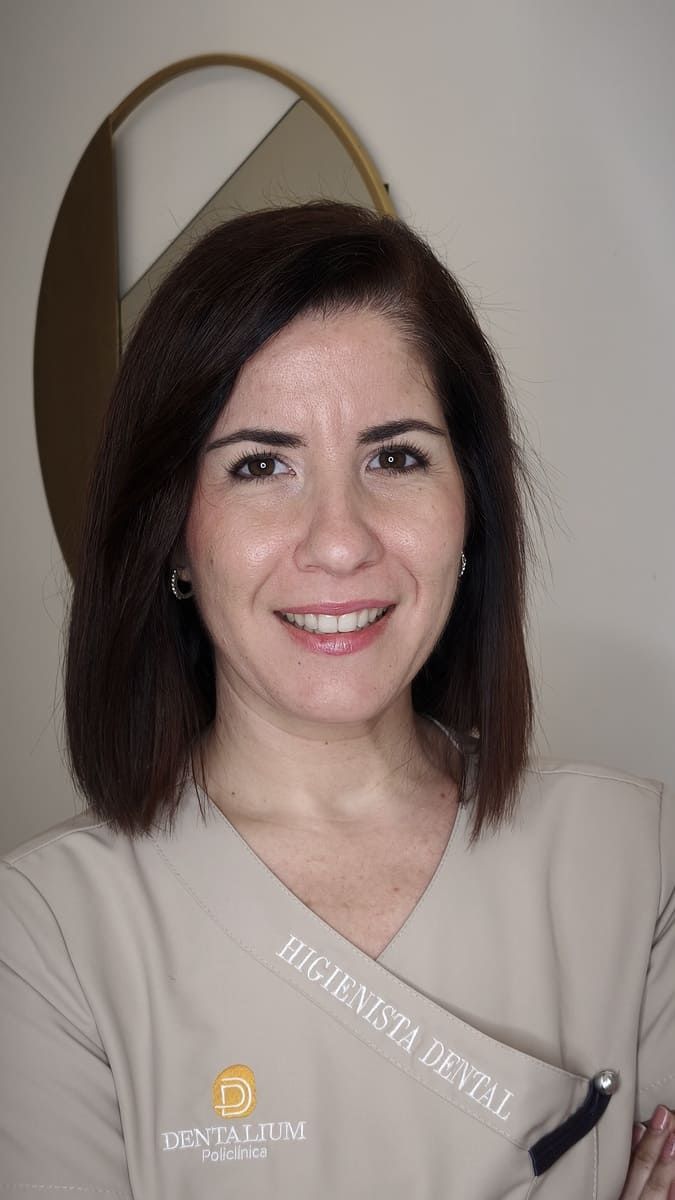 Mujer sonriente con camisa de uniforme color beige, apoyada contra una pared clara, con un espejo decorativo detrás de ella.