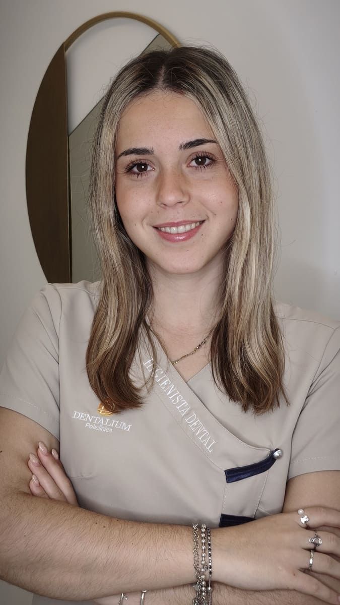 Persona sonriente con cabello largo y una blusa beige, sentada contra una pared lisa con un arco decorativo detrás.