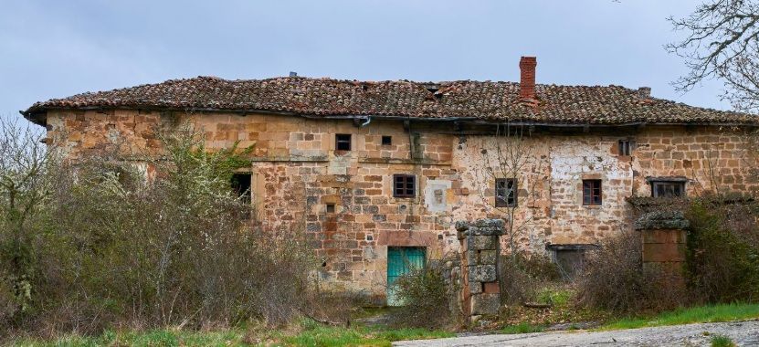 Edificio antiguo de piedra erosionada por el tiempo, con tejado de tejas de arcilla. Puerta verde y arbustos frondosos en la entrada.