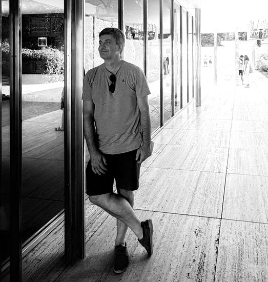 Un hombre en pantalones cortos y camiseta se apoya en una columna, con las piernas cruzadas, afuera de un edificio moderno.