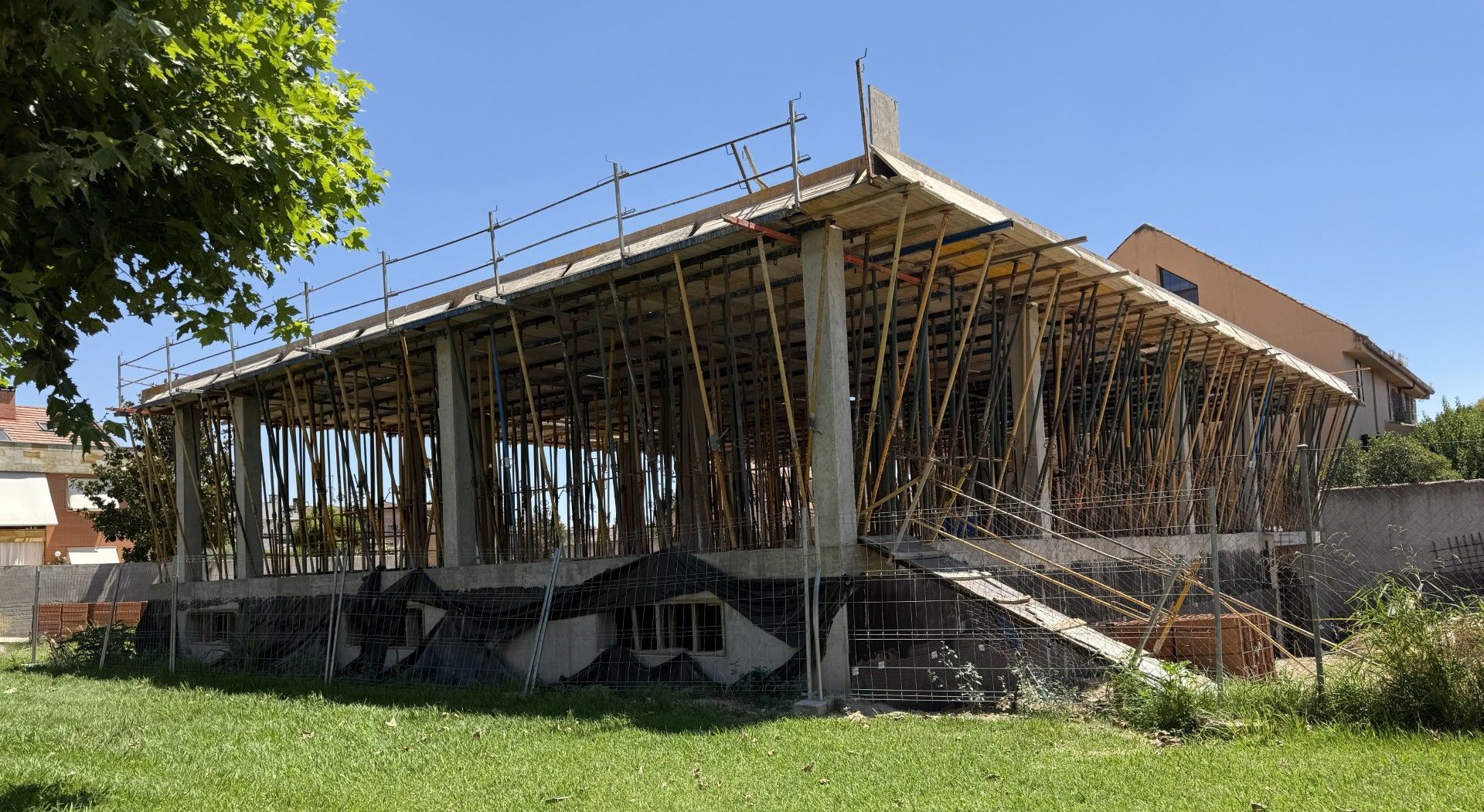 Edificio en construcción, estructura de hormigón sostenida por andamios sobre un césped, fondo de cielo azul.