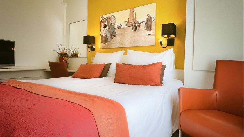 Hotelkamer met oranje accenten: bed, kussens, kunstwerken, stoelen en een gele muur.
