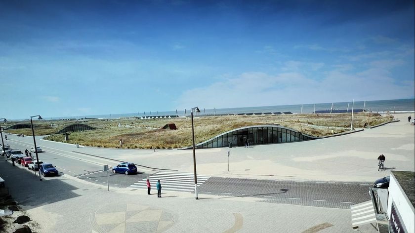 Een strandtafereel met een gebogen grasveld, voetgangers, een blauwe lucht en auto's.