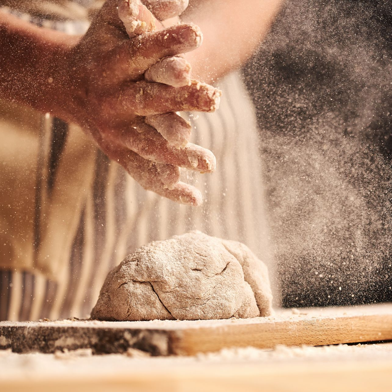 Des mains avec de la farine pétrissent la pâte sur une planche en bois, dans une cuisine chaleureuse.