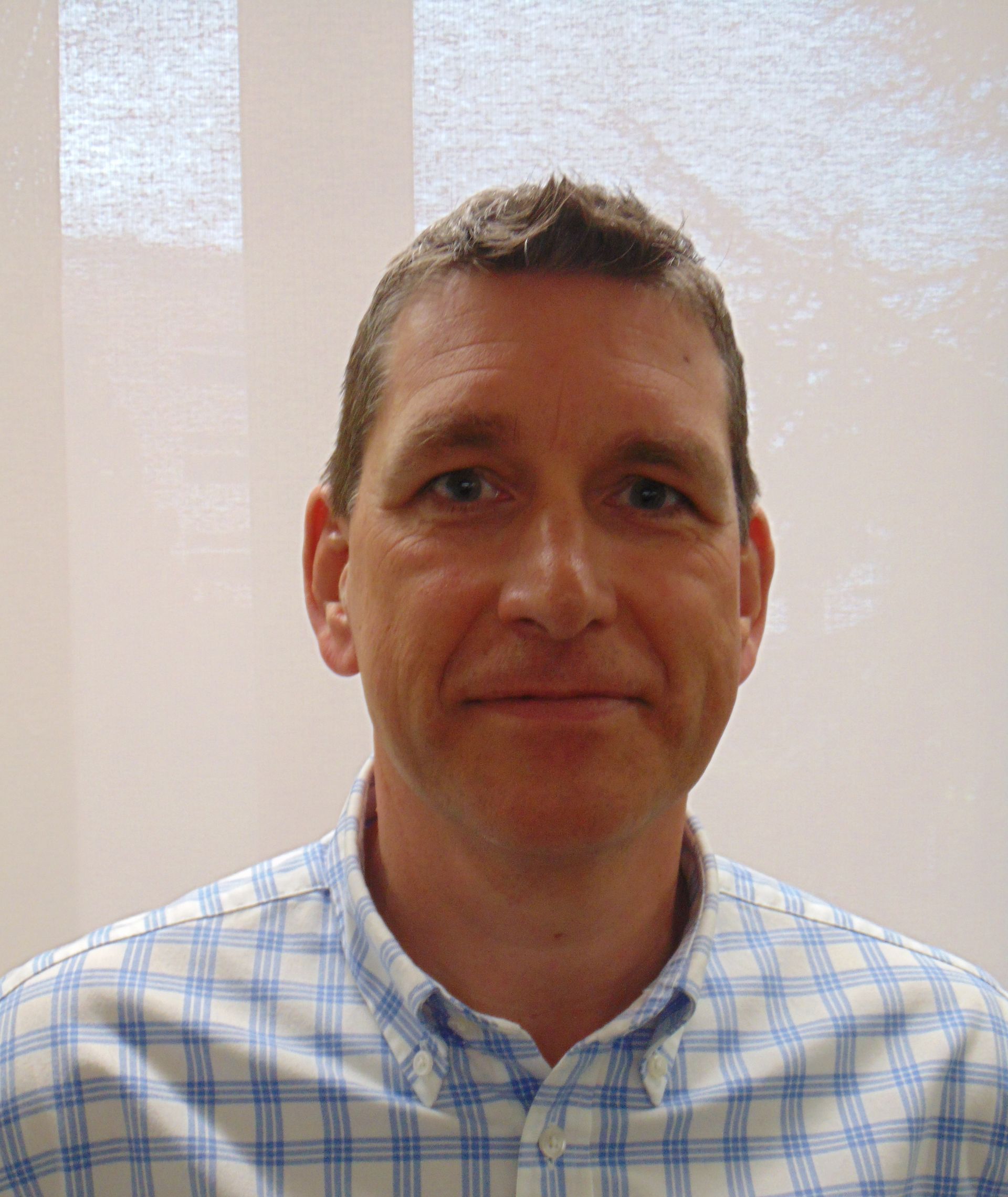 Mann in hellblauem kariertem Hemd lächelt; neutraler Hintergrund.