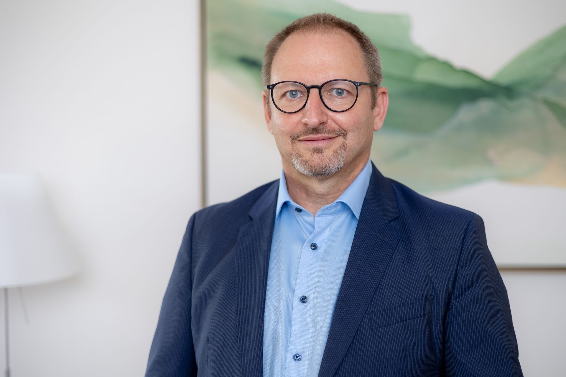 Ein Mann in Anzug und Brille steht vor einem Gemälde.