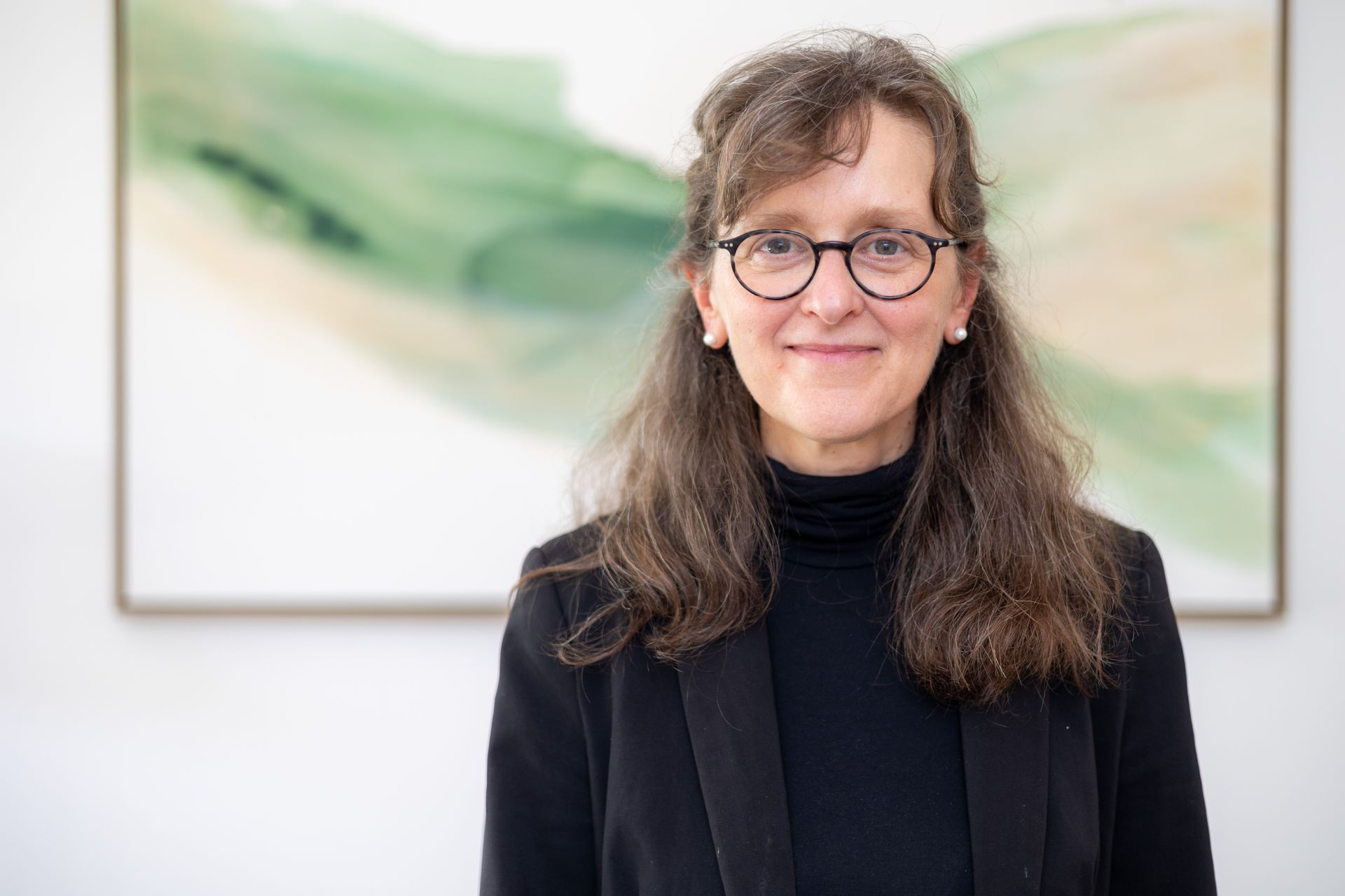 Eine Frau mit Brille und schwarzer Jacke steht vor einem Gemälde.