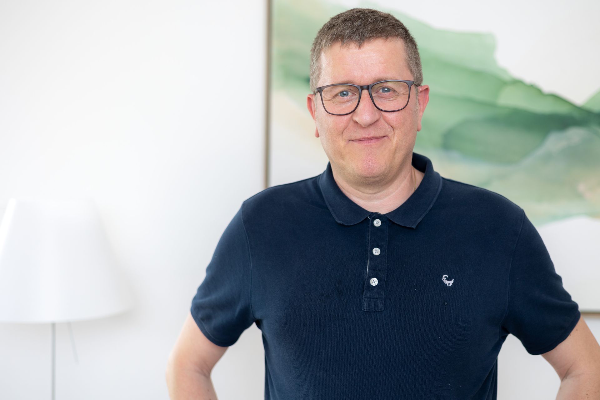 Ein Mann mit Brille und blauem Poloshirt steht vor einem Gemälde.
