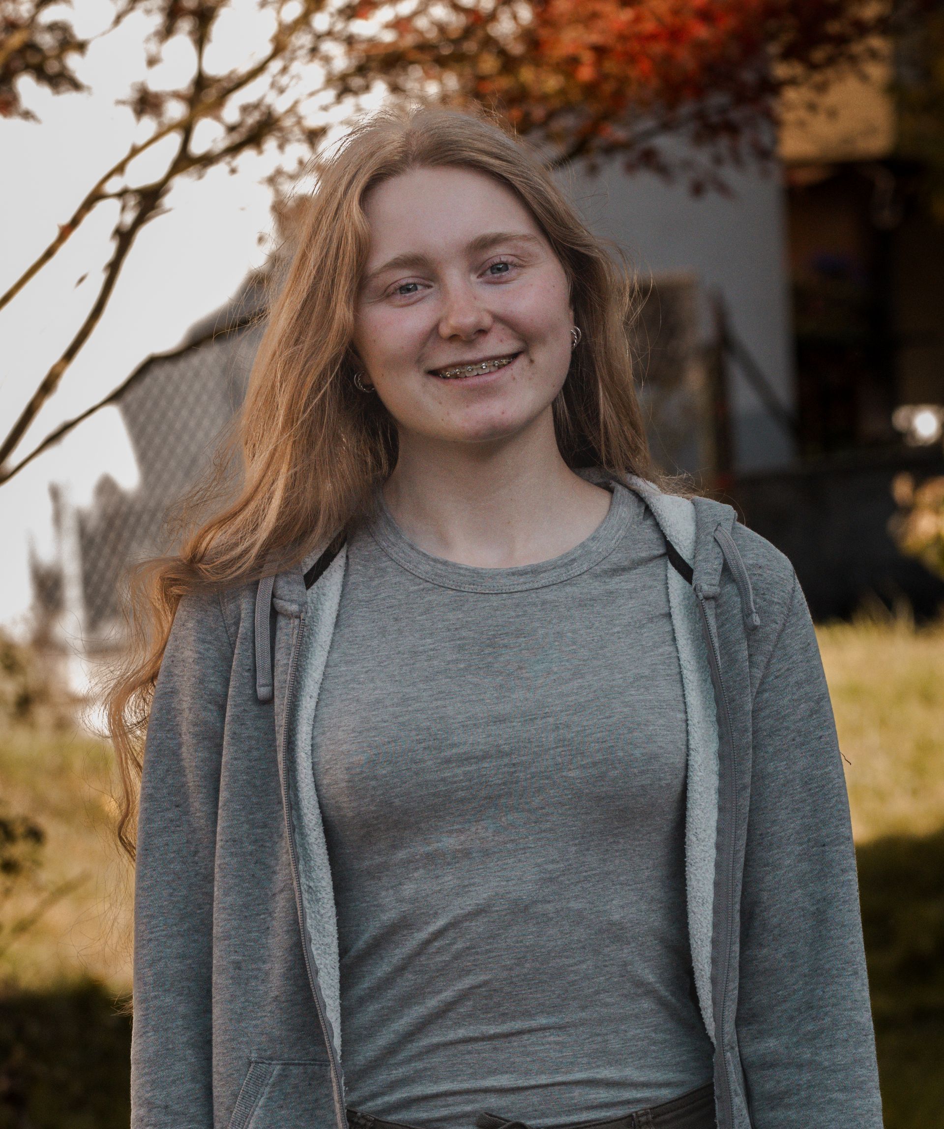 Eine junge Frau in einem grauen Shirt und einem grauen Kapuzenpullover steht vor einem Baum.