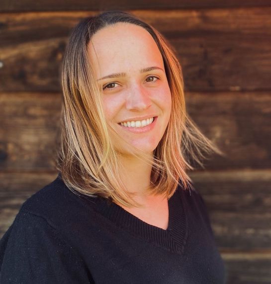 Eine Frau in einem schwarzen Pullover lächelt vor einer Holzwand.