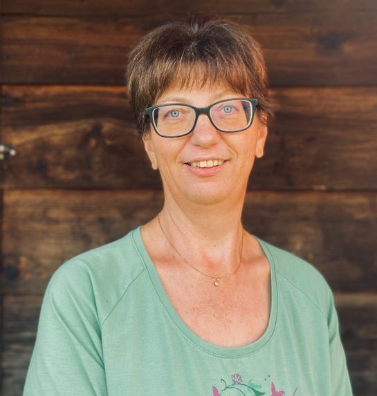 Eine Frau mit Brille und grünem Hemd lächelt vor einer Holzwand.