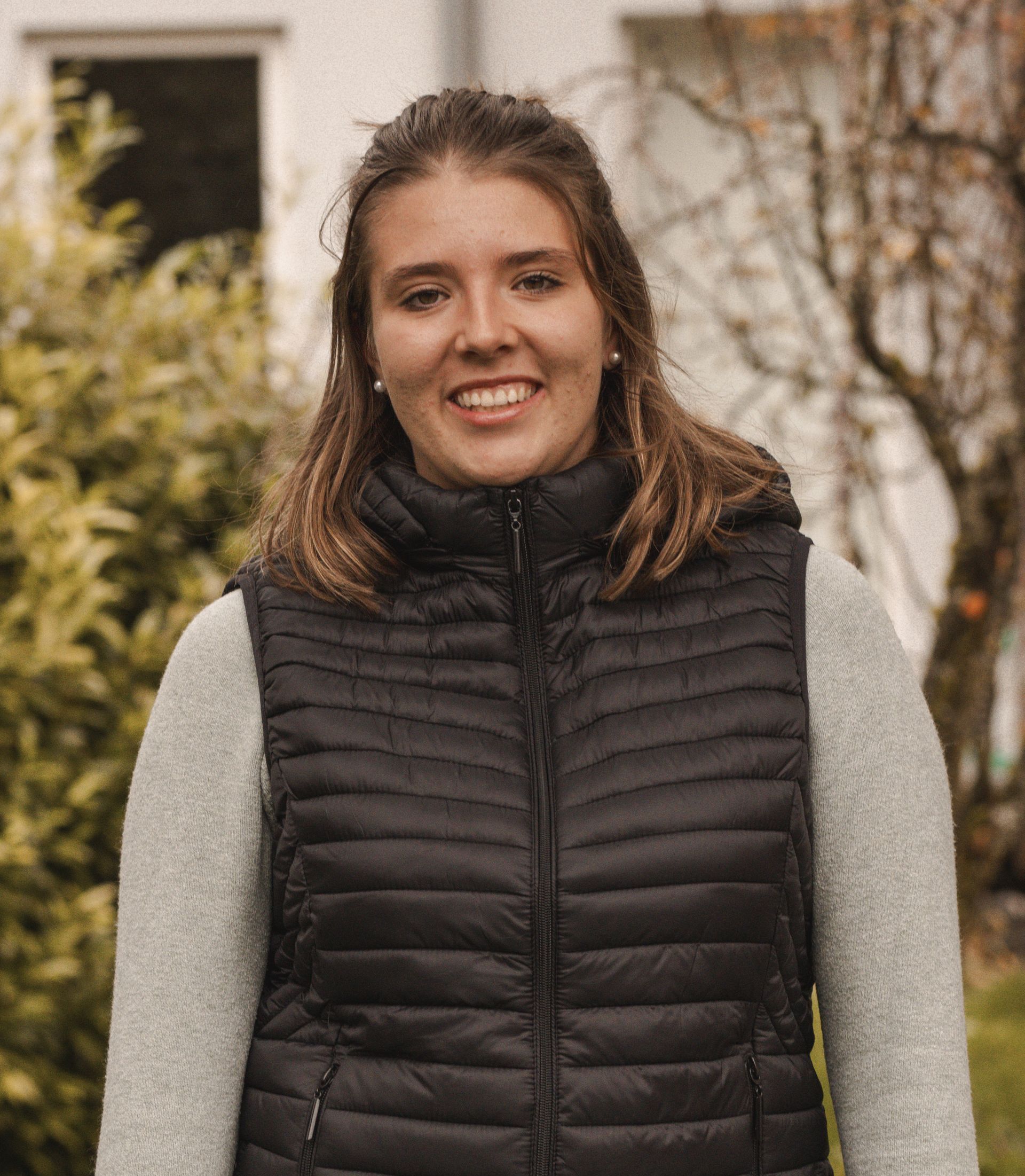 Eine Frau mit einer schwarzen Weste und einem grauen Pullover steht vor einem Haus.