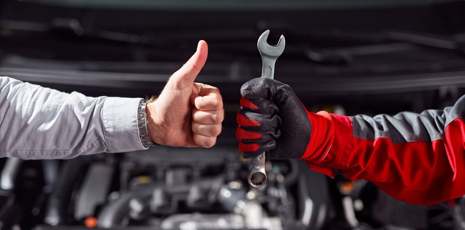Ein Mann zeigt mit dem Daumen nach oben und hält dabei einen Schraubenschlüssel vor einem Auto.