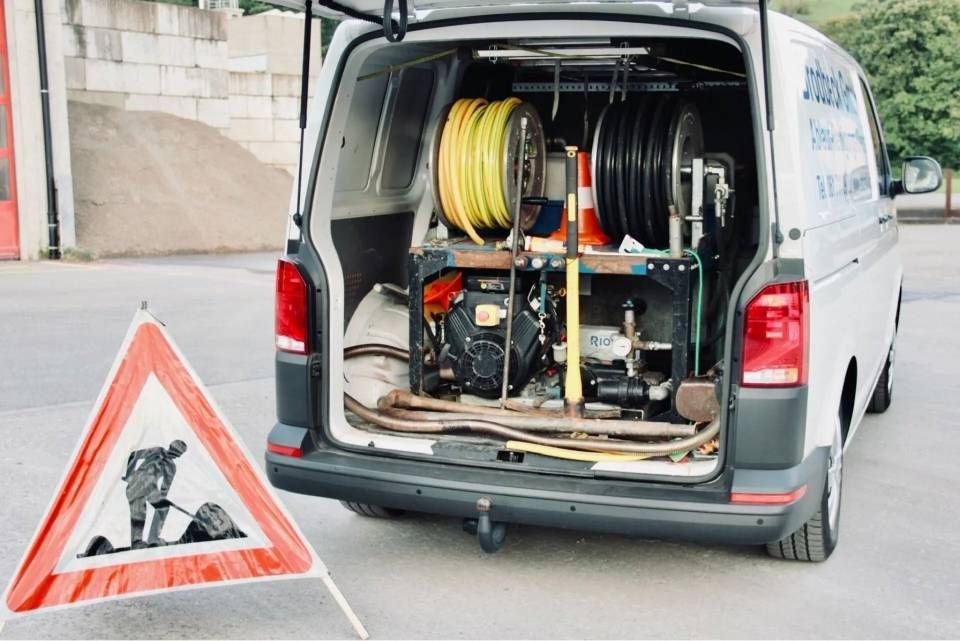 Weisser Lieferwagen mit geöffneten Hecktüren, aus dem Ausrüstung und Schläuche zu sehen sind, neben einem Warnschild für eine Strassenbaustelle