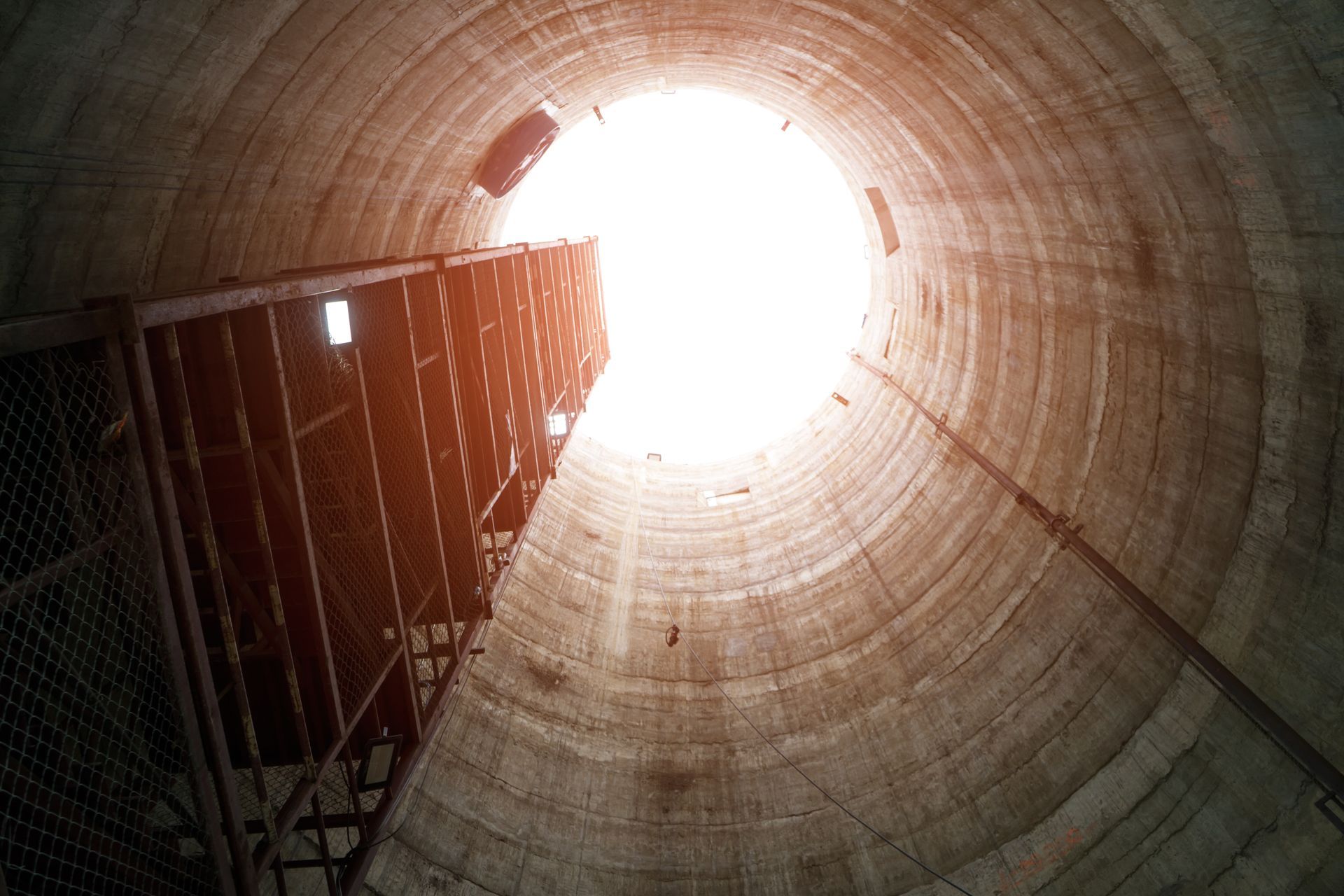 Der Blick schweift in das Innere eines hohen, zylindrischen Silos zu einer hellen Öffnung hinauf, links davon befindet sich eine verrostete Metalltreppe.