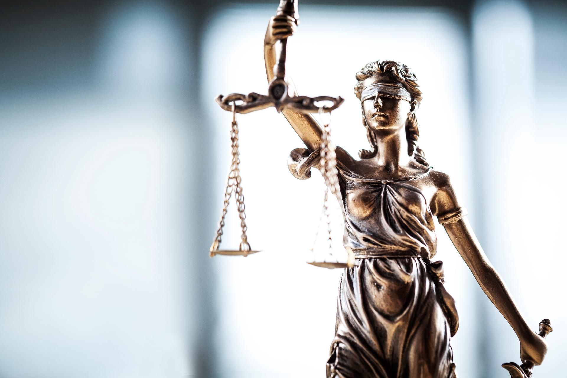 Eine Bronzestatue der Justitia mit einer Waage in der Hand vor einem sanften, hellen Hintergrund.