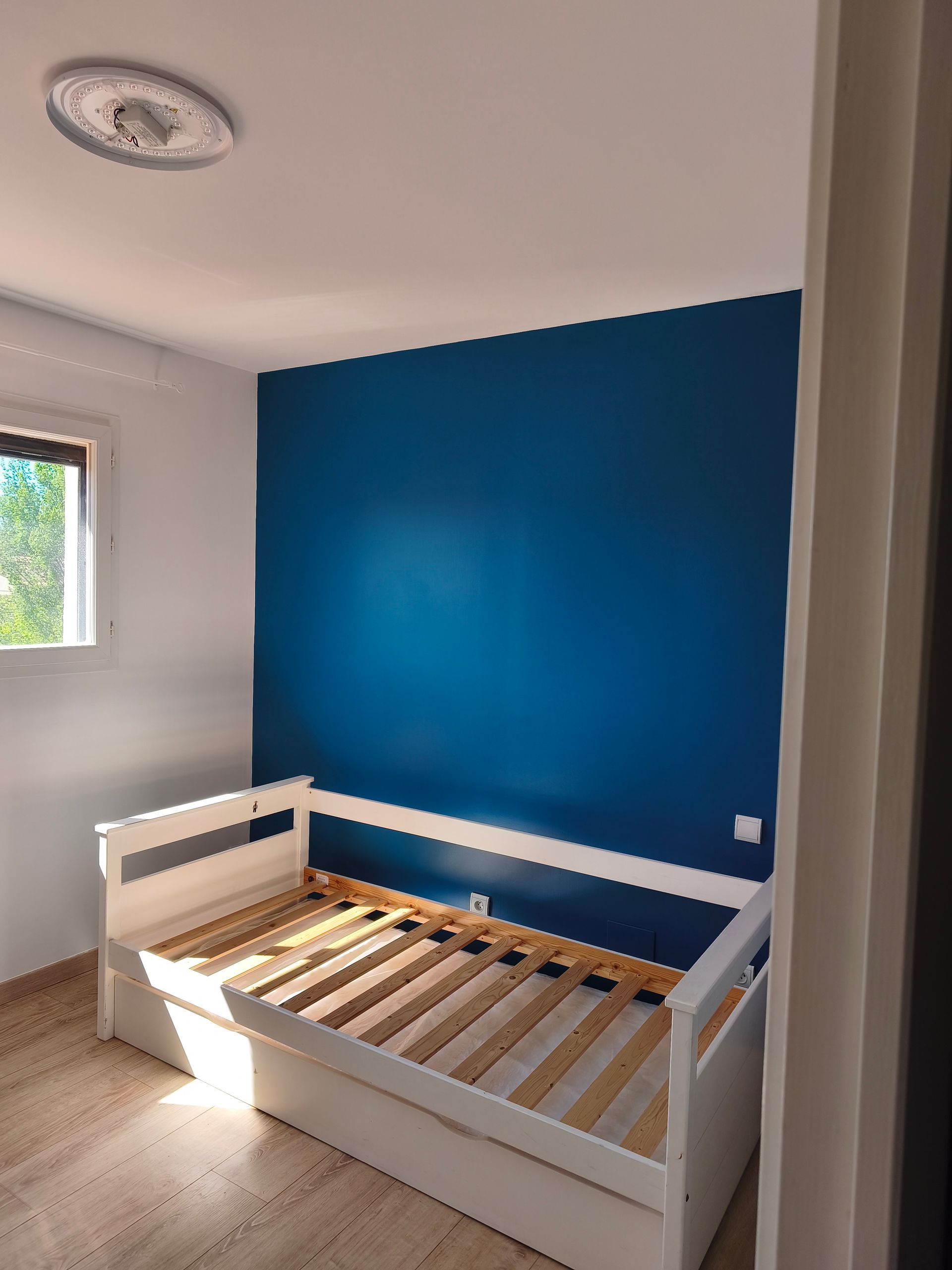 Chambre vide avec un mur bleu, un cadre de lit blanc et une petite fenêtre.