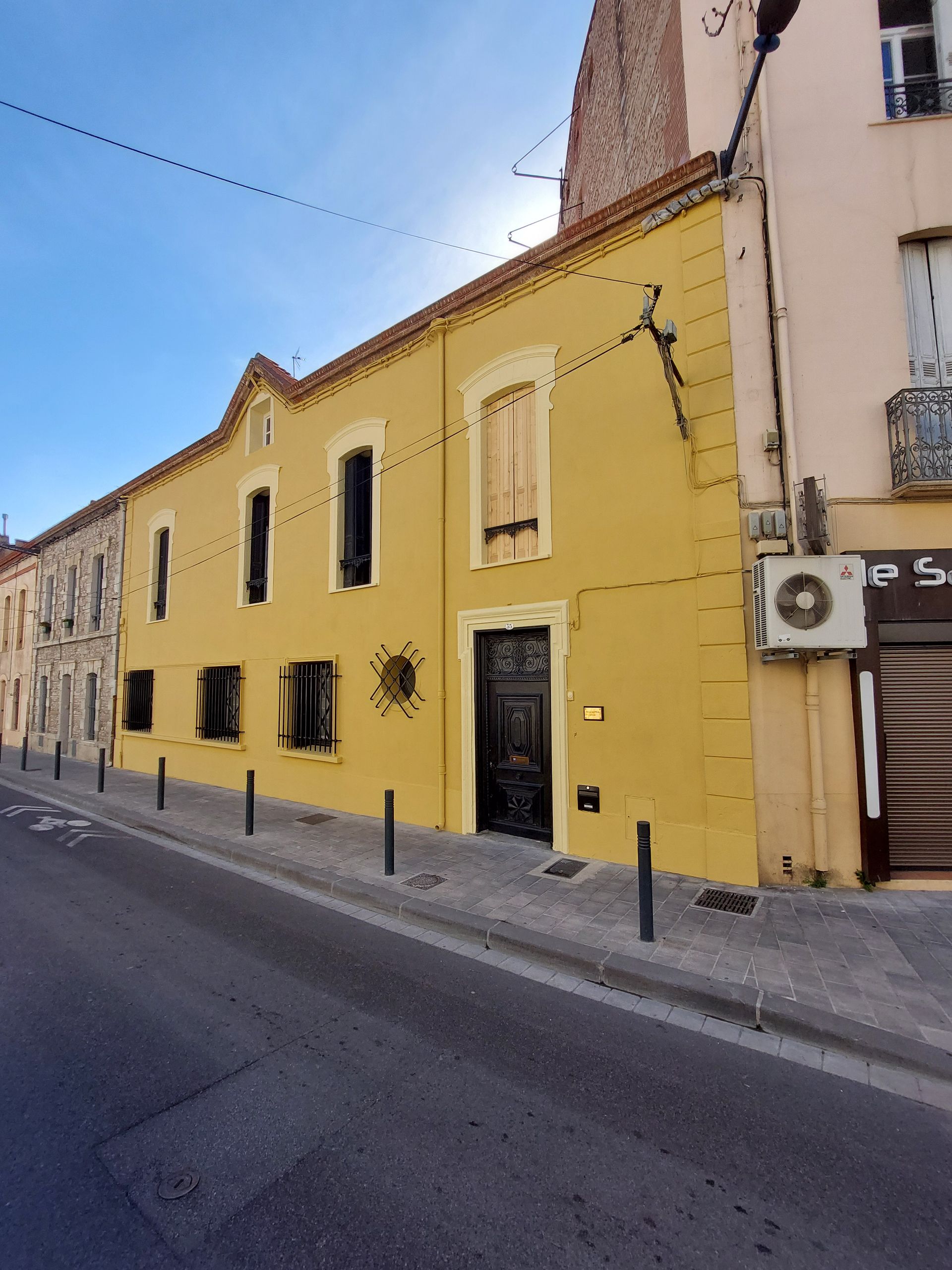 Bâtiment en stuc jaune dans une rue calme, avec des fenêtres à volets et une porte sous un ciel bleu.
