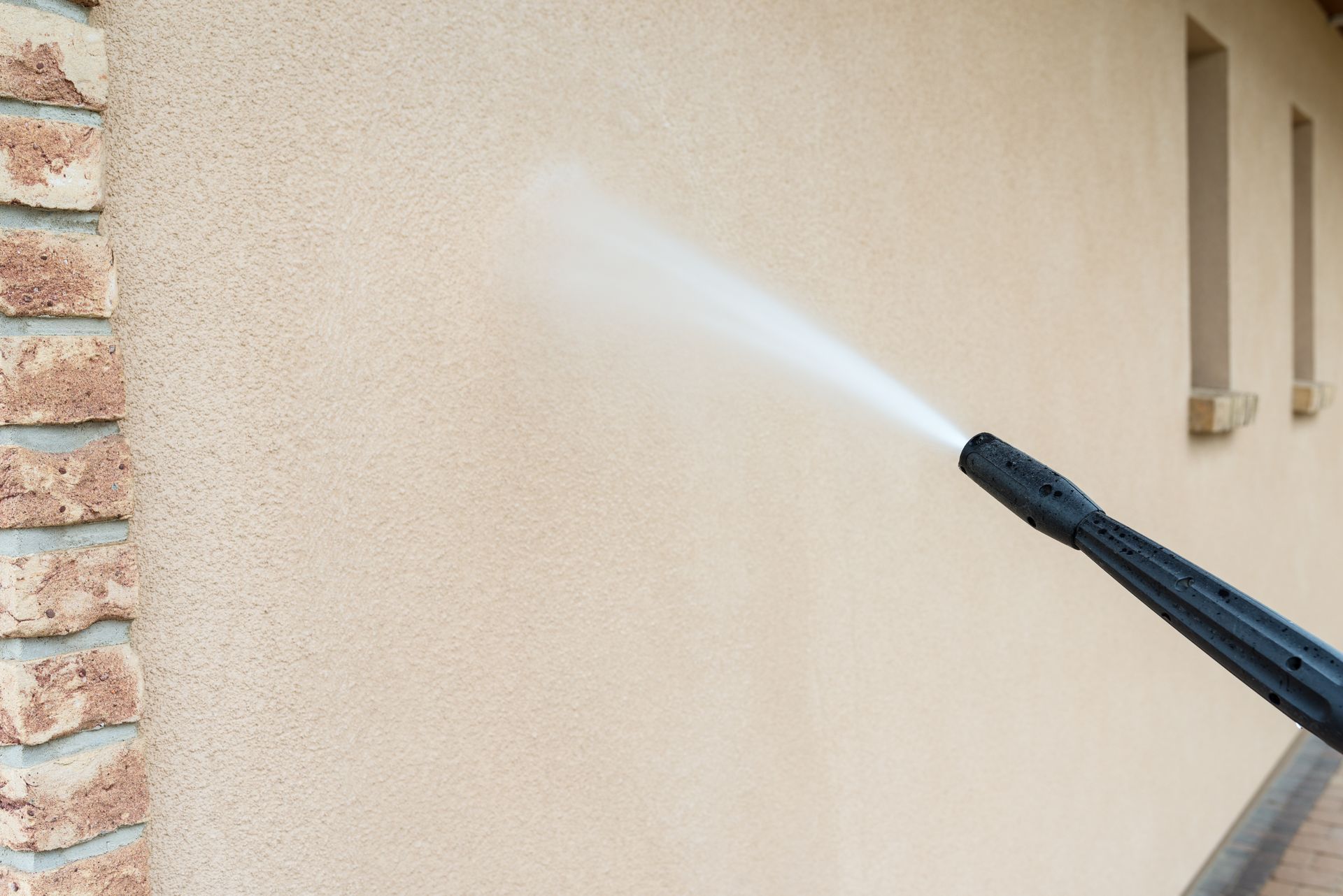 La buse d'un nettoyeur à haute pression projette un fin brouillard d'eau contre le mur d'une maison en stuc beige.
