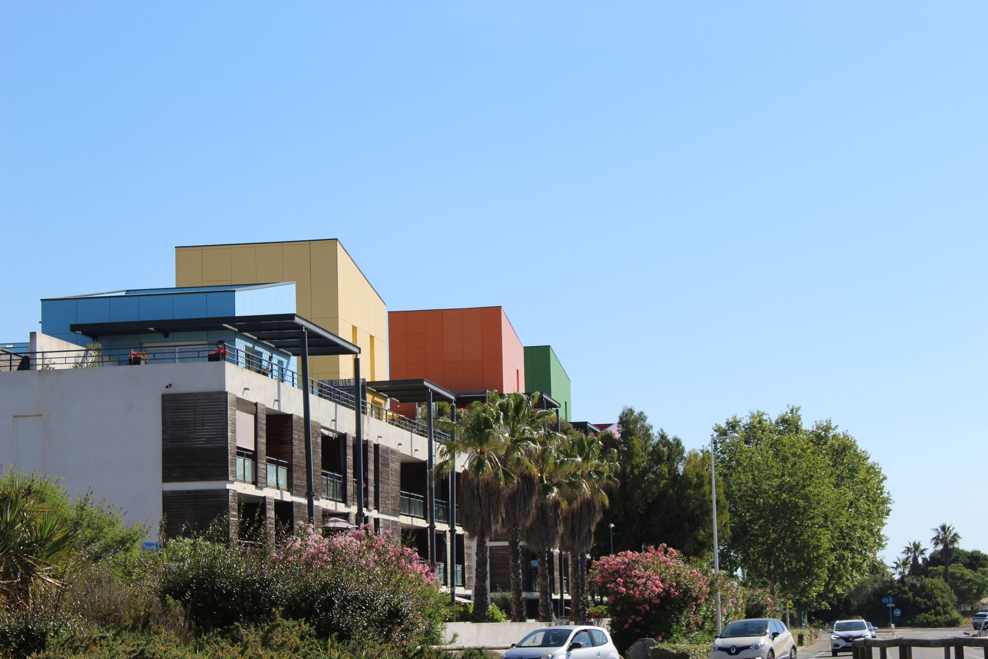 Bâtiment moderne aux toits colorés et aux palmiers, en bordure d'une route sous un ciel d'un bleu limpide.