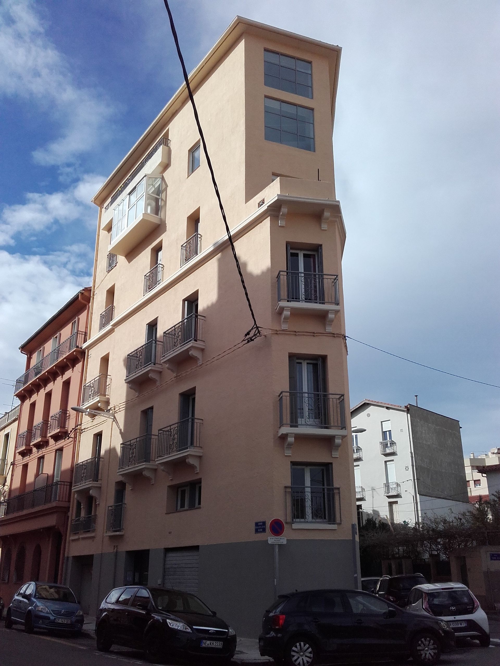 Un immeuble étroit, en forme de coin, avec une façade rose pâle, se dresse à un coin de rue sous un ciel bleu nuageux.