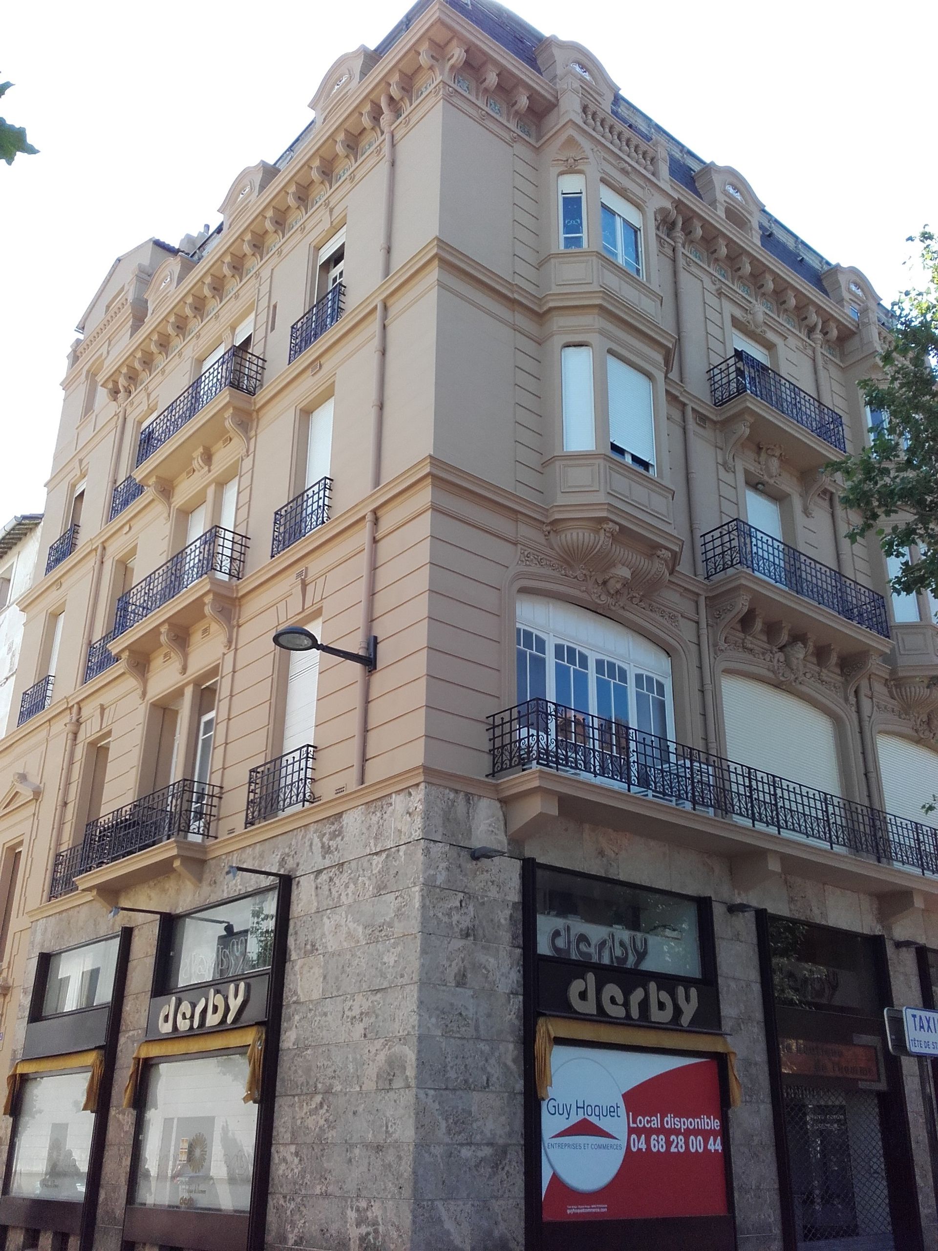 Façade d'un immeuble d'angle beige à plusieurs étages, avec un soubassement en pierre