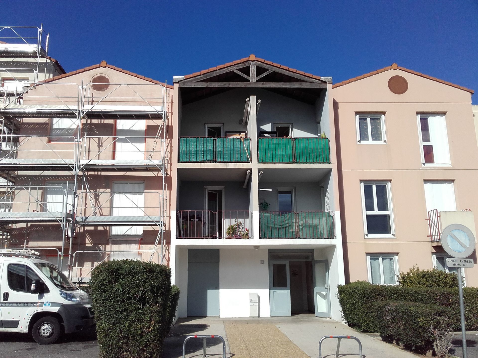 Immeuble d'appartements de trois étages avec balcons et véhicules stationnés dans une rue ensoleillée