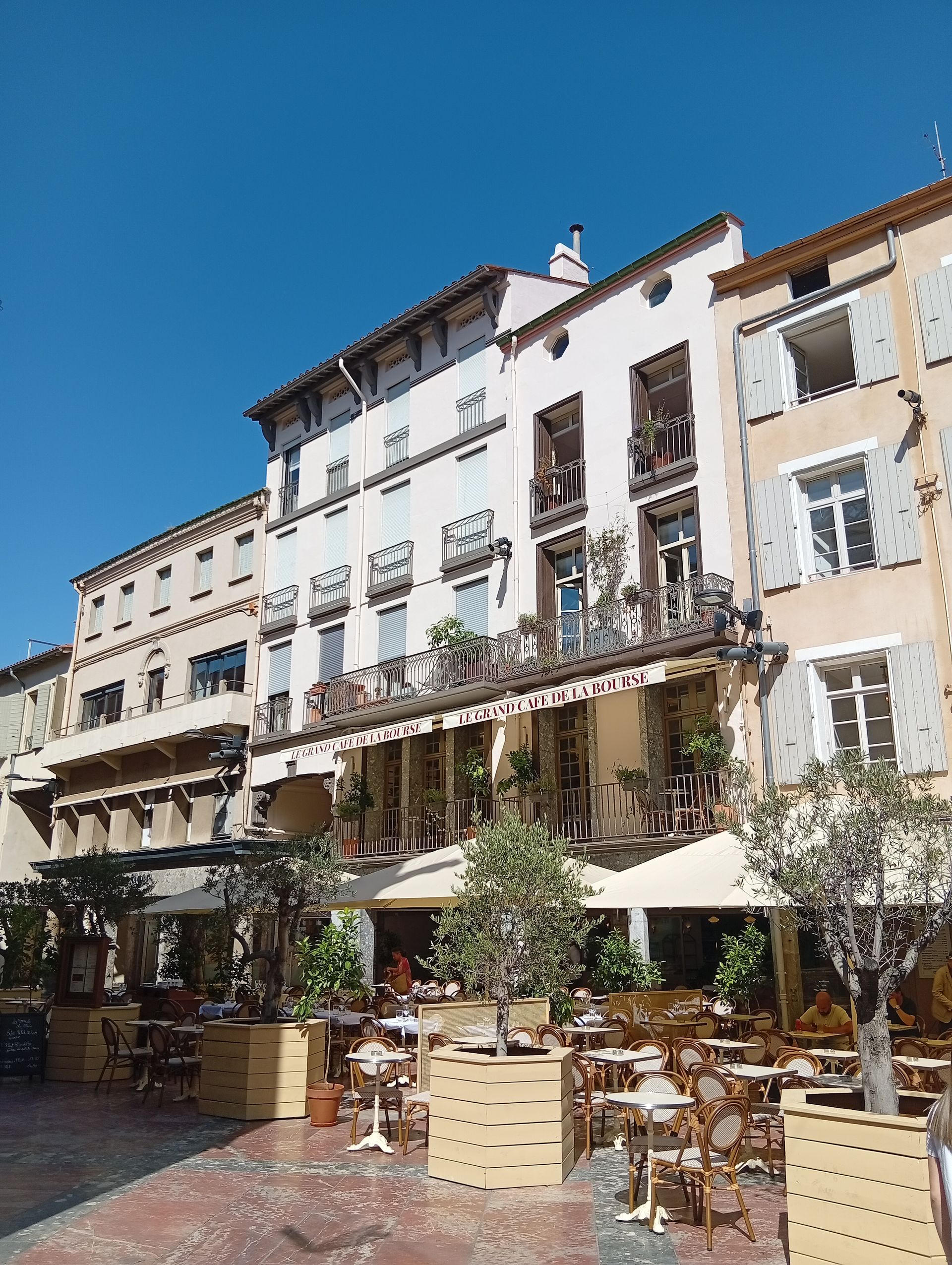 Terrasse ensoleillée d'un café avec parasols et tables devant un bâtiment historique blanc sur une place de ville