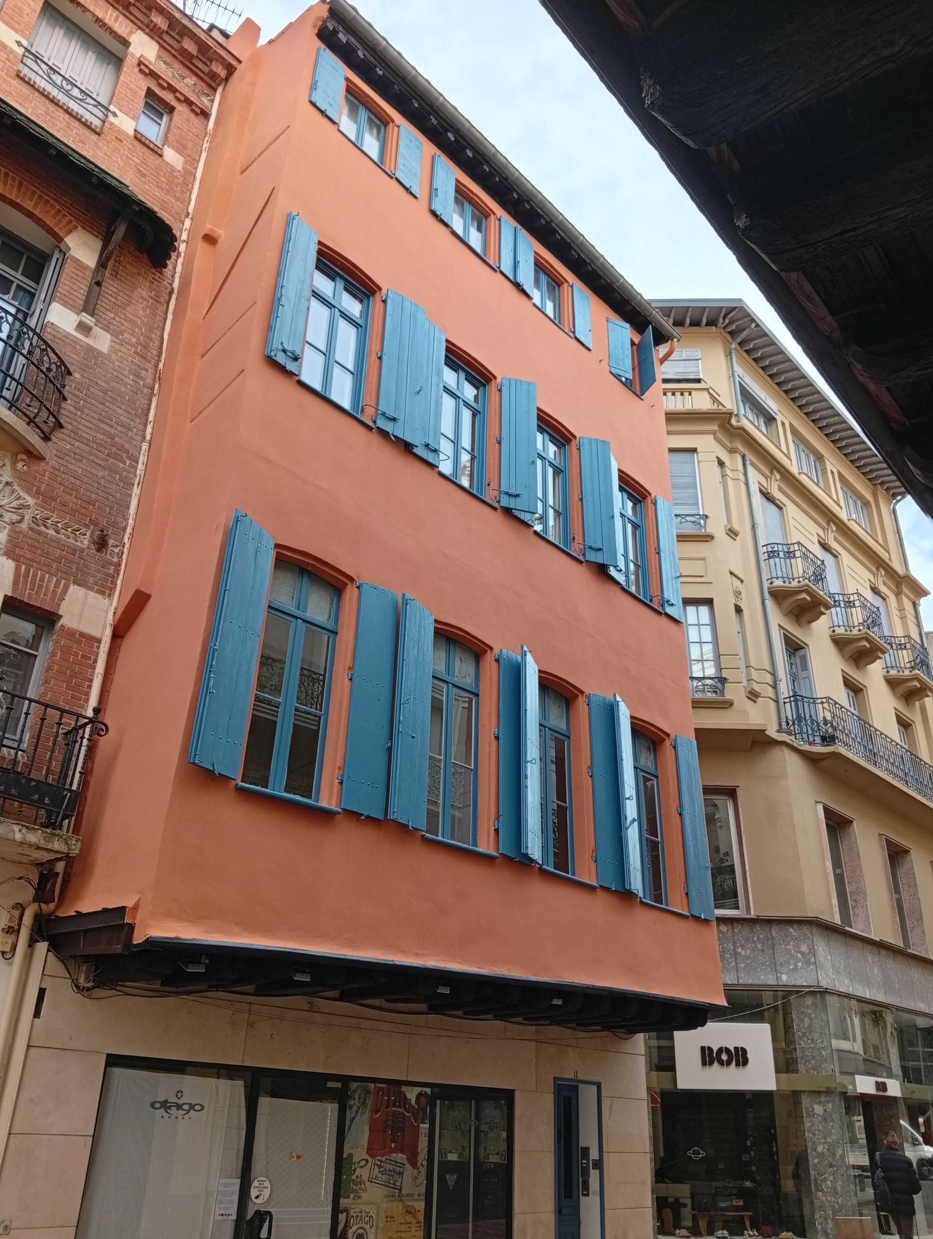 Rue étroite bordée d'un immeuble corail aux volets bleus, à côté de bâtiments beiges plus anciens.