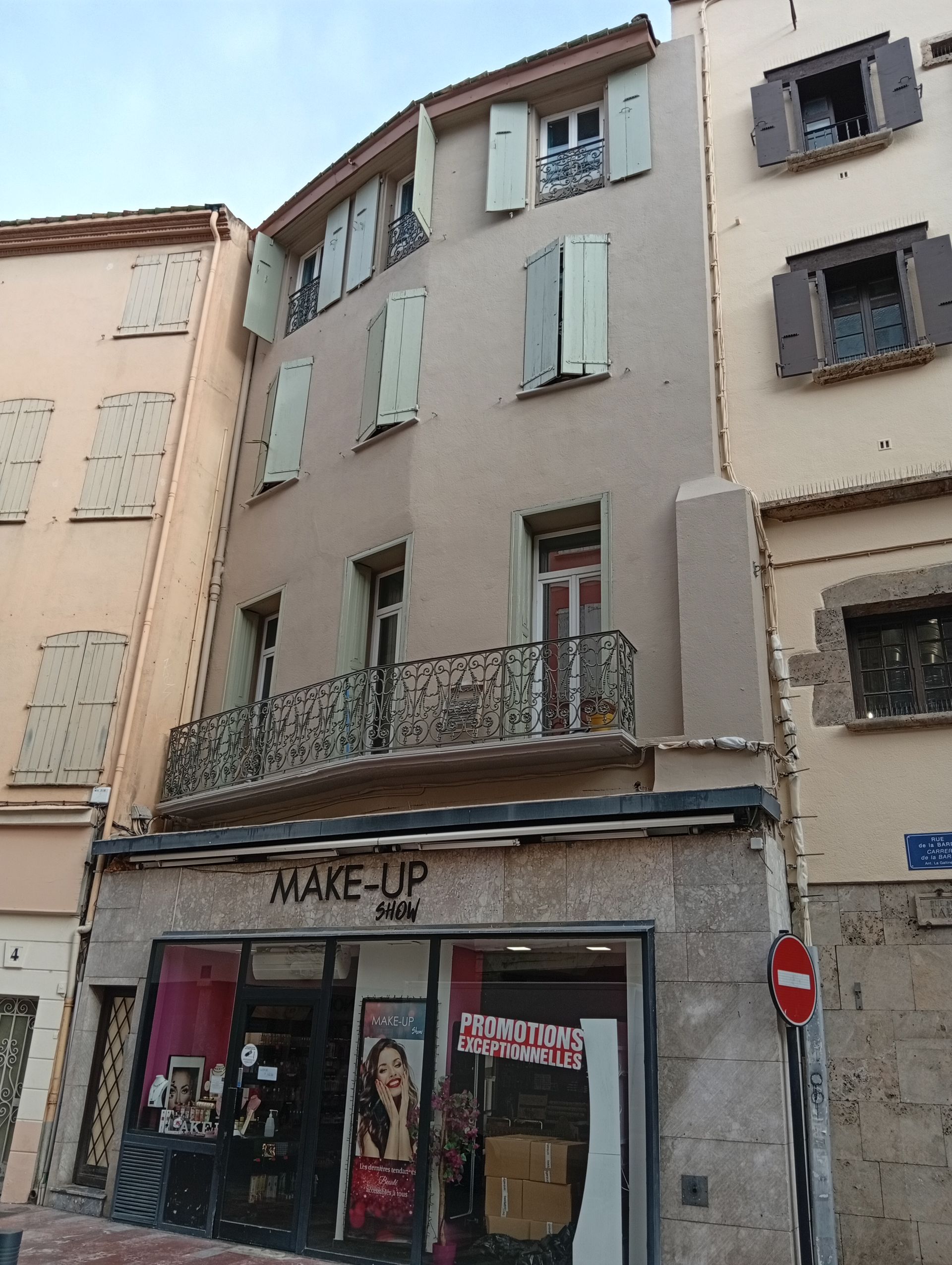 Façade de magasin européenne étroite sous des appartements en stuc usé, avec des volets verts et des balcons.