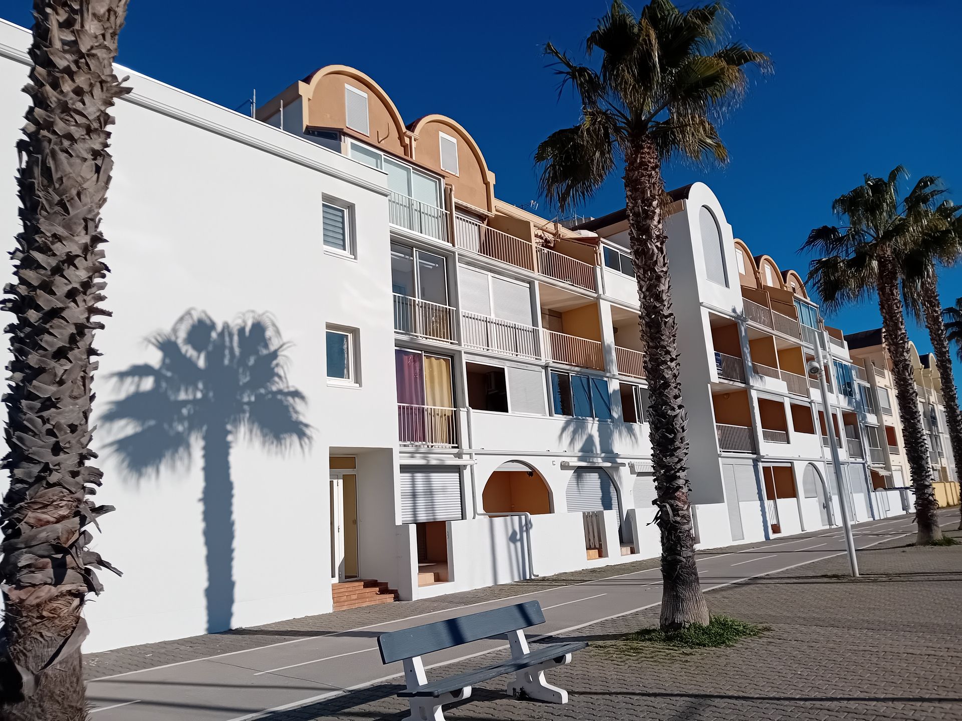 Immeuble d'appartements blanc baigné de soleil, avec balcons, palmiers et un banc devant