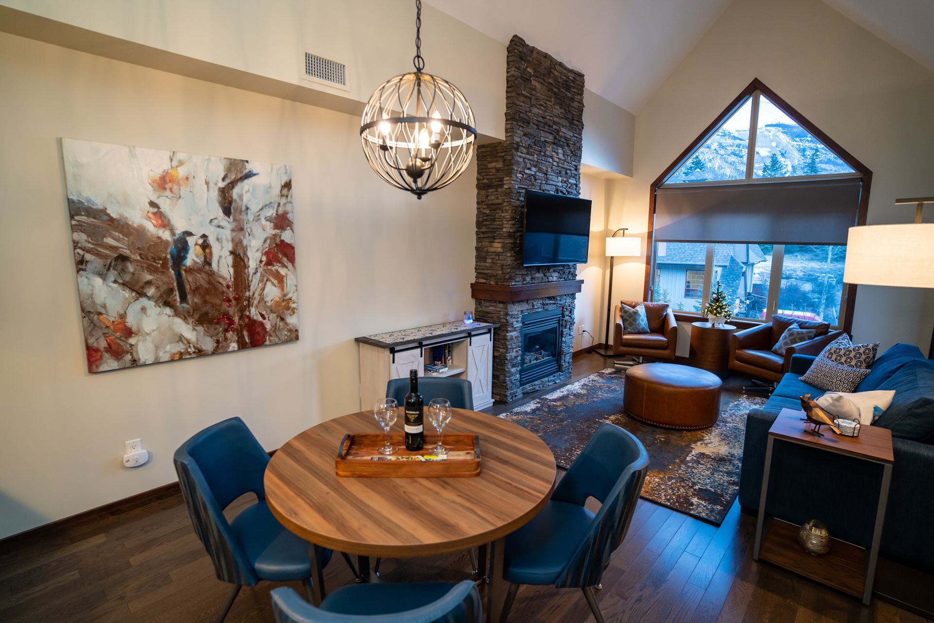 A modern cabin living room: dining table, fireplace, artwork, seating, and a large window.