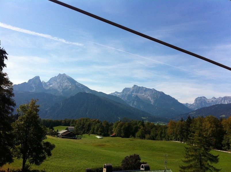 Berglandschaft mit grünen Feldern, vereinzelten Bäumen und einem blauen Himmel mit zarten Wolken