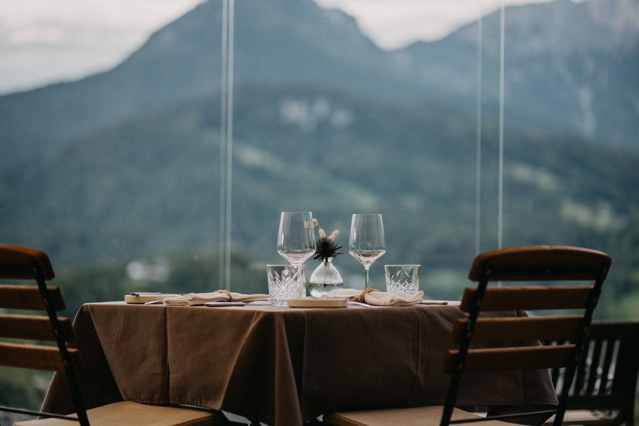 Restauranttisch mit Weingläsern, von dem aus man durch große Fenster einen verschwommenen Blick auf die Berge hat.