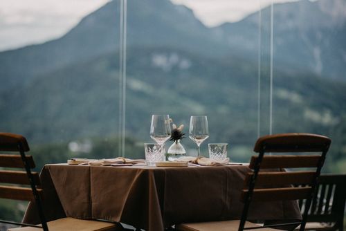 Restauranttisch mit Weingläsern, von dem aus man durch große Fenster einen verschwommenen Blick auf die Berge hat.