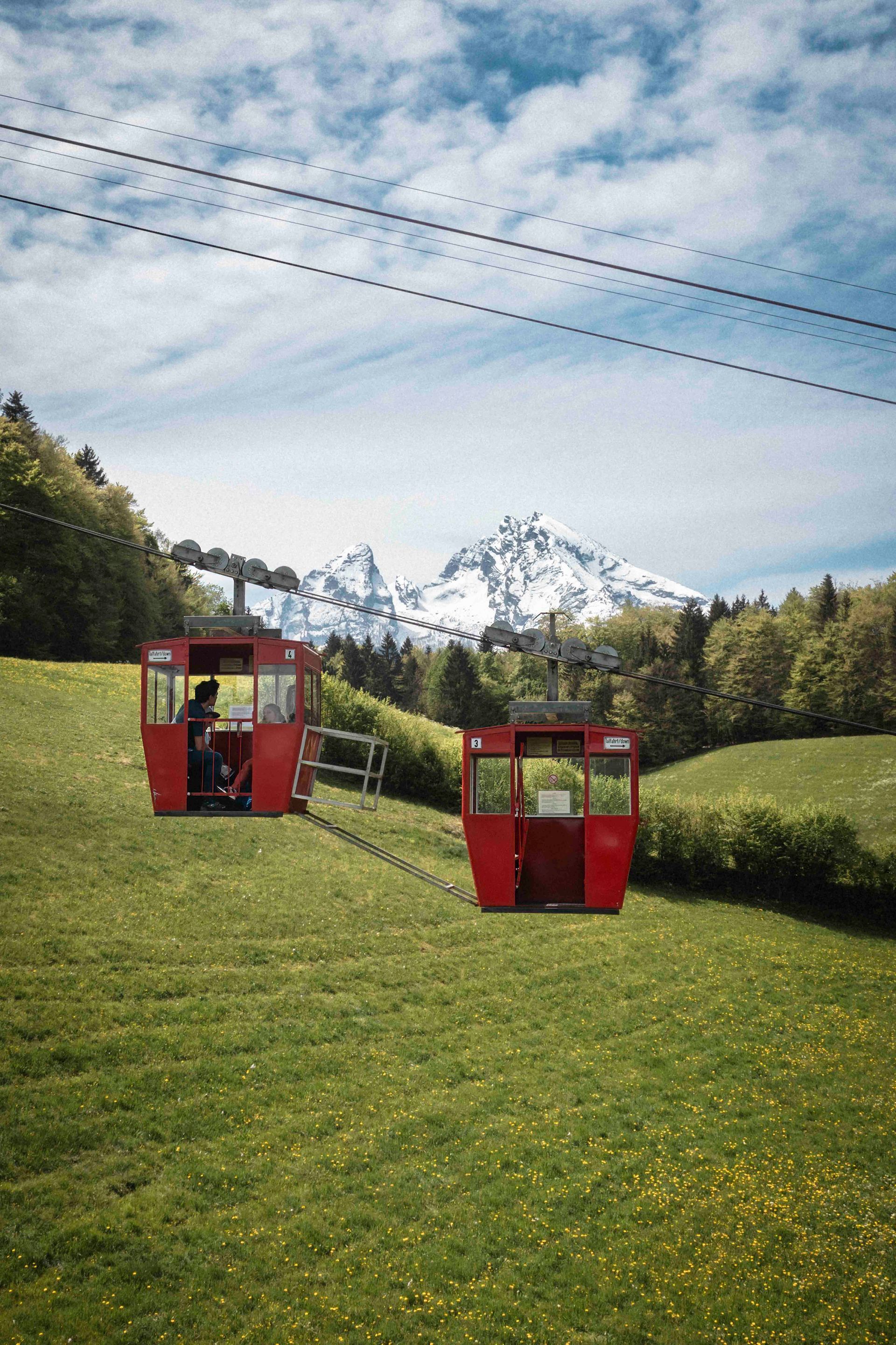 Zwei rote Seilbahnwagen auf einem grasbewachsenen Hang vor dem Hintergrund schneebedeckter Berge und blauem Himmel.