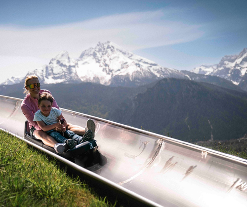 Zwei Personen fahren auf einem silbernen Rodel einen Hang hinunter, im Hintergrund erheben sich schneebedeckte Berge.