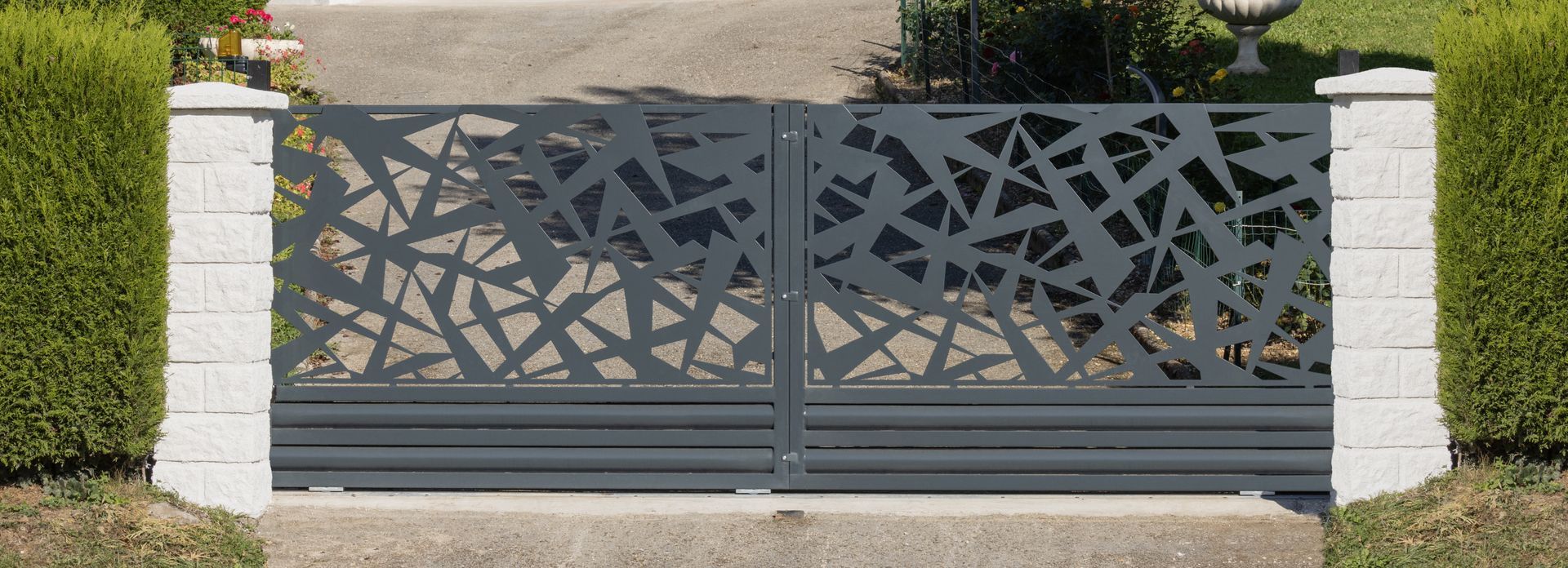 Portail d'allée moderne en métal gris foncé à motifs géométriques, entre des piliers en pierre blanche.