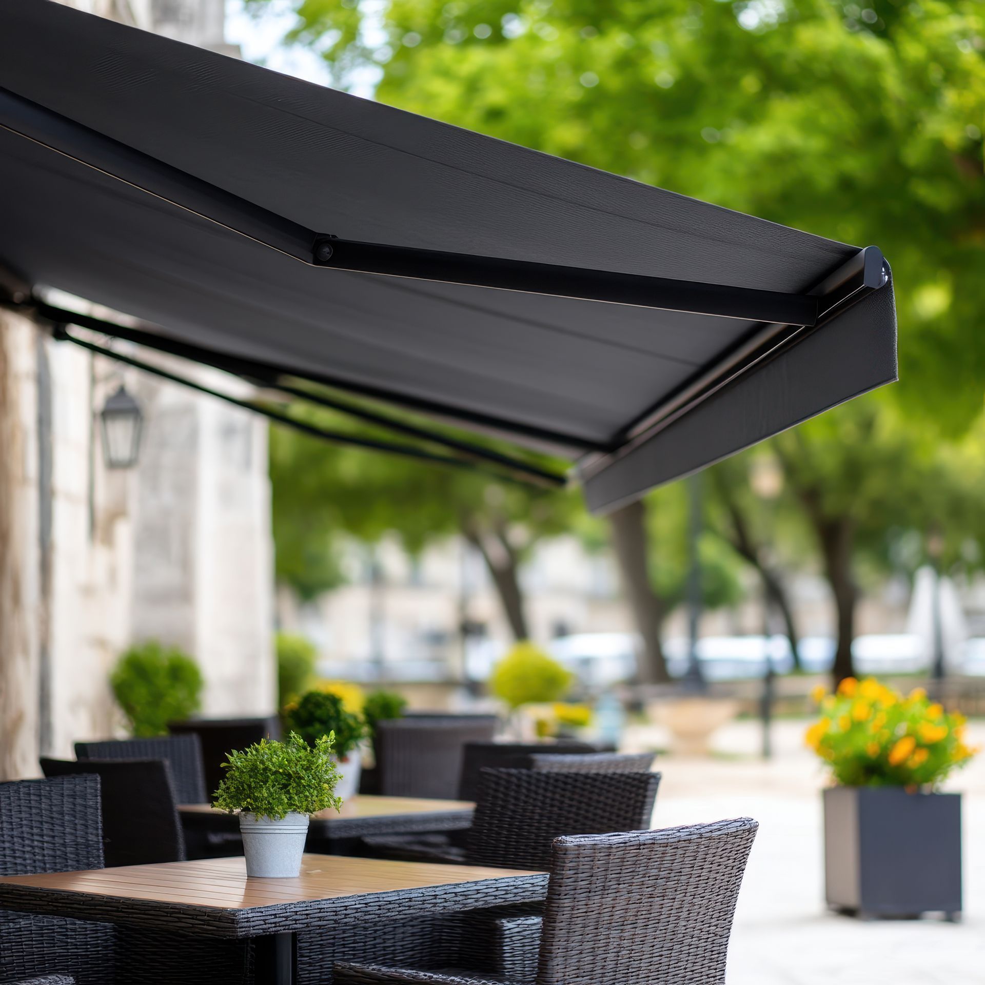 Terrasse de café sous un auvent noir, avec tables et chaises en osier, plantes en pot et arbres verts en arrière-plan.