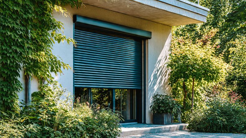 Entrée d'une maison moderne avec un volet métallique bleu fermé, encadrée par une végétation luxuriante de lierre.