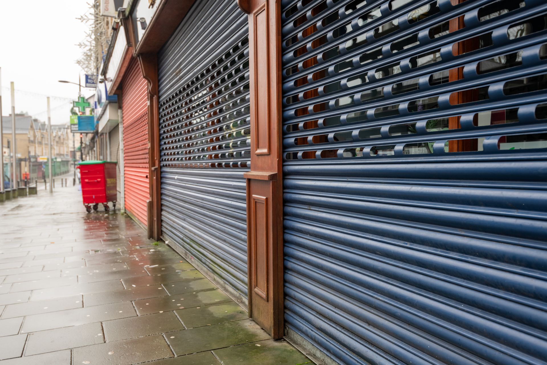 Rue pluvieuse, rideaux métalliques de magasins fermés et trottoir mouillé.