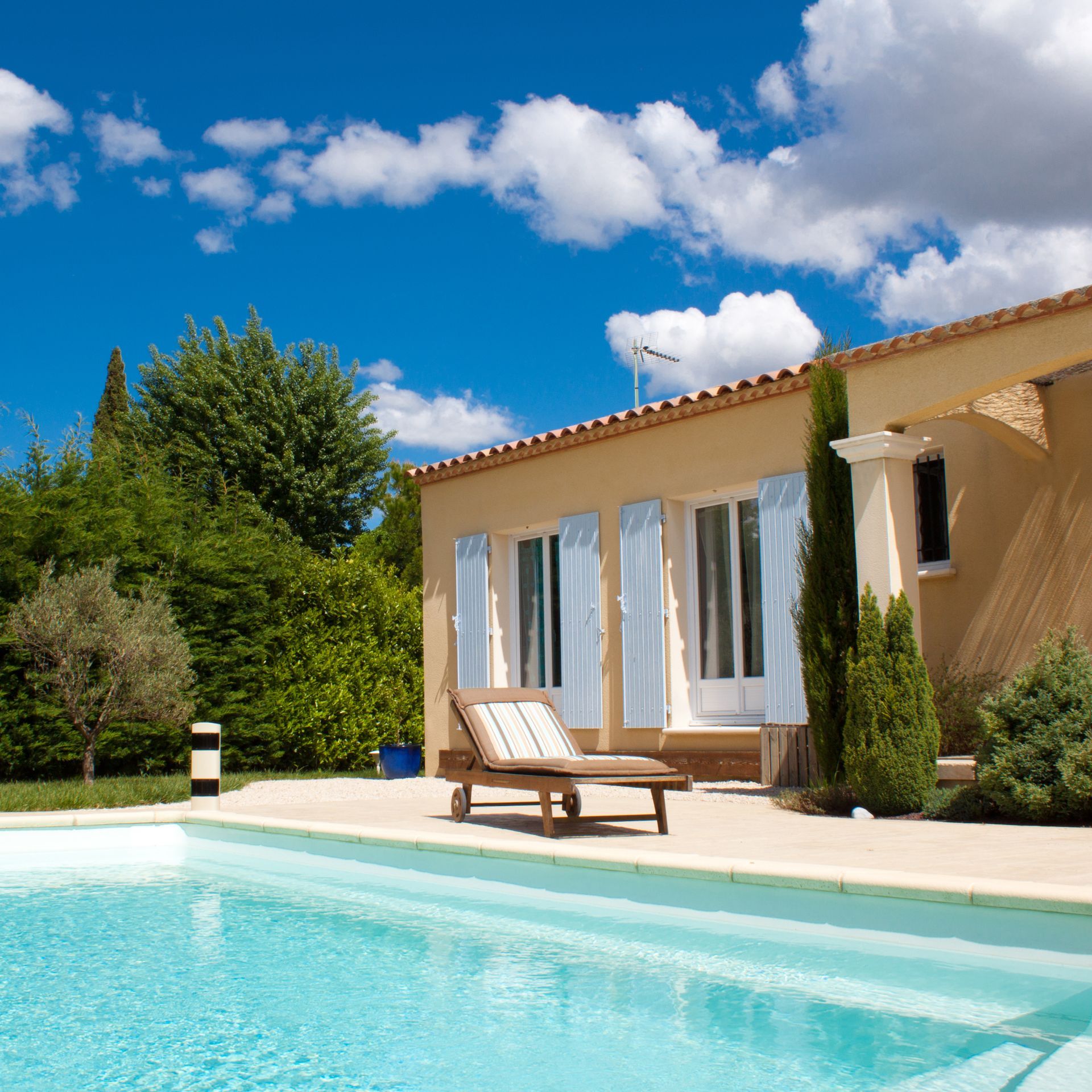 Maison moderne avec piscine turquoise, chaise longue sur la terrasse et ciel bleu parsemé de nuages.
