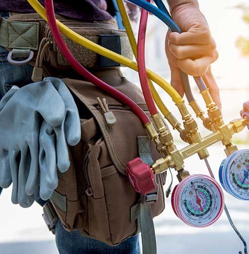 Técnico de HVAC sosteniendo medidores con mangueras, usando cinturón de herramientas y guantes de trabajo al aire libre.