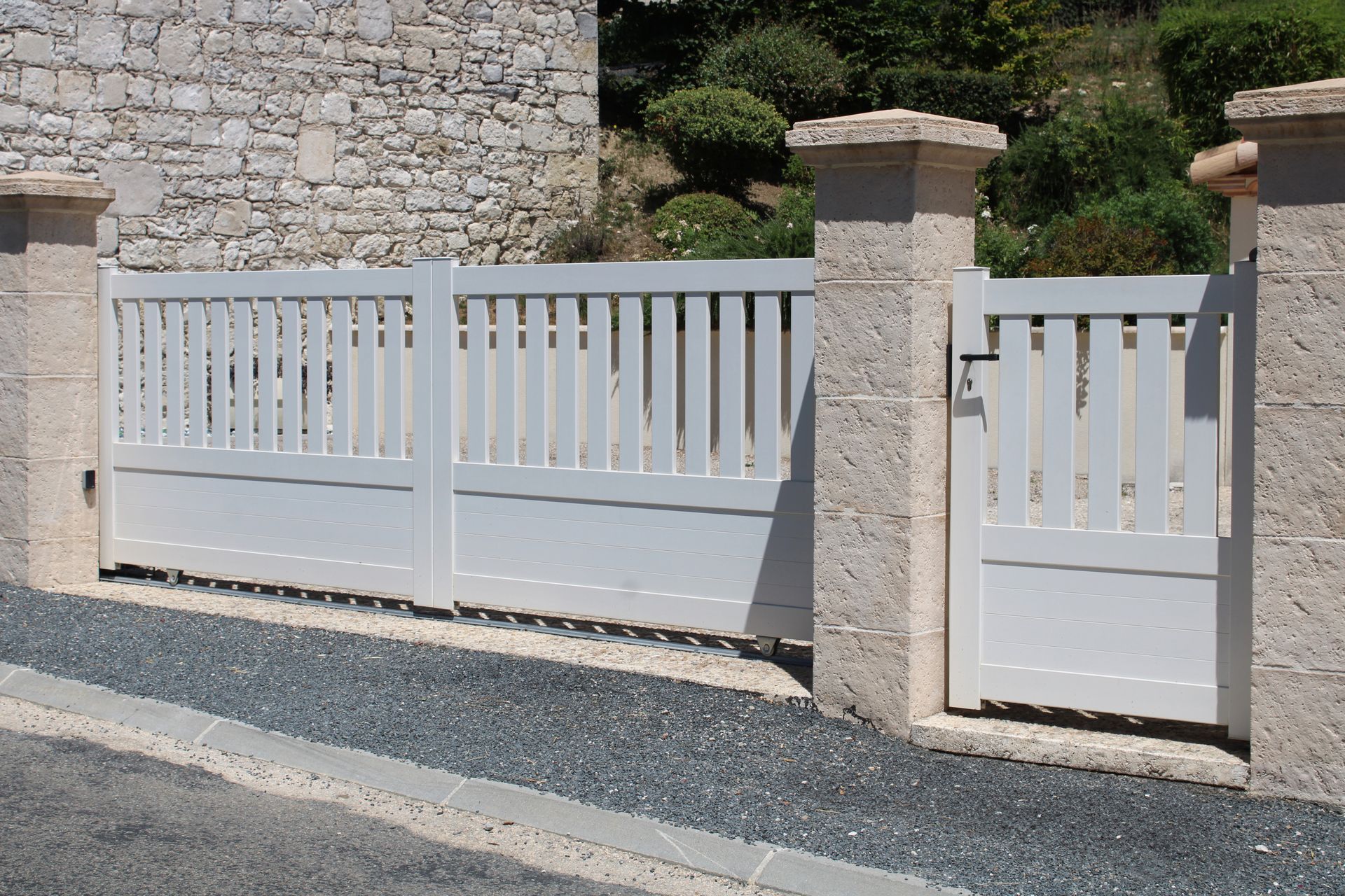 Entrée blanche avec portail, barreaux verticaux et piliers en pierre. Allée en gravier, muret en pierre en arrière-plan.