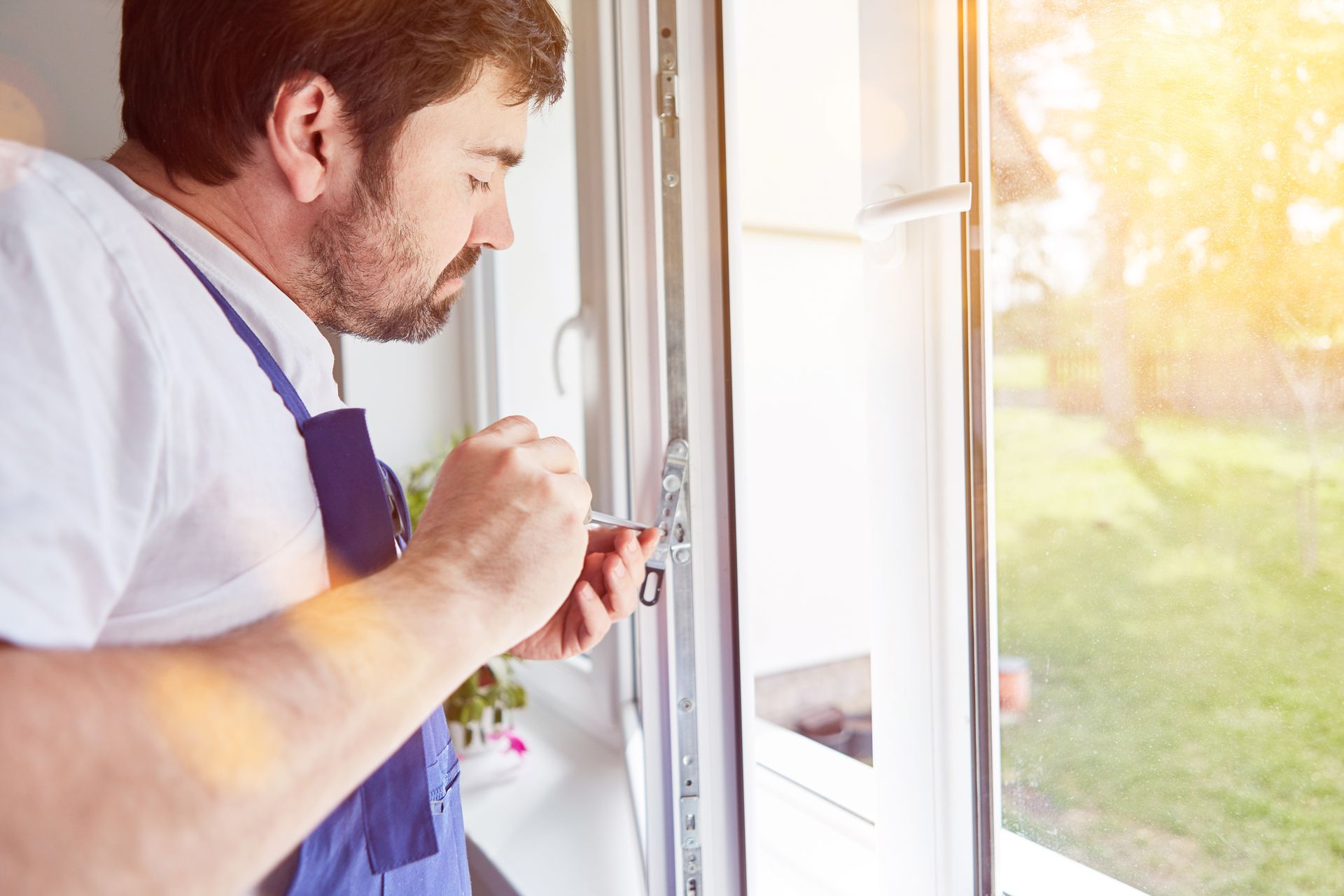 Un homme en chemise blanche et tablier bleu répare une fenêtre avec un tournevis, tandis que la lumière du soleil entre.