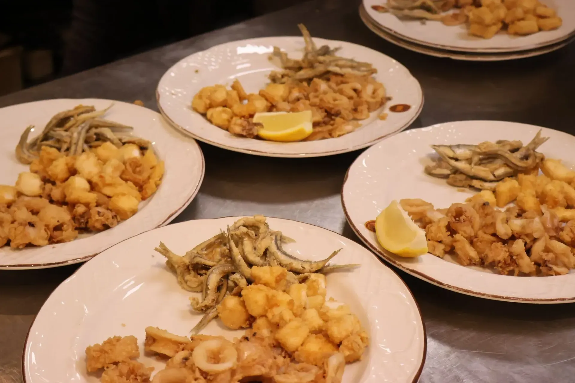 Varios platos blancos sobre un mostrador, cada uno con porciones de marisco frito, aros de calamar y una rodaja de limón.