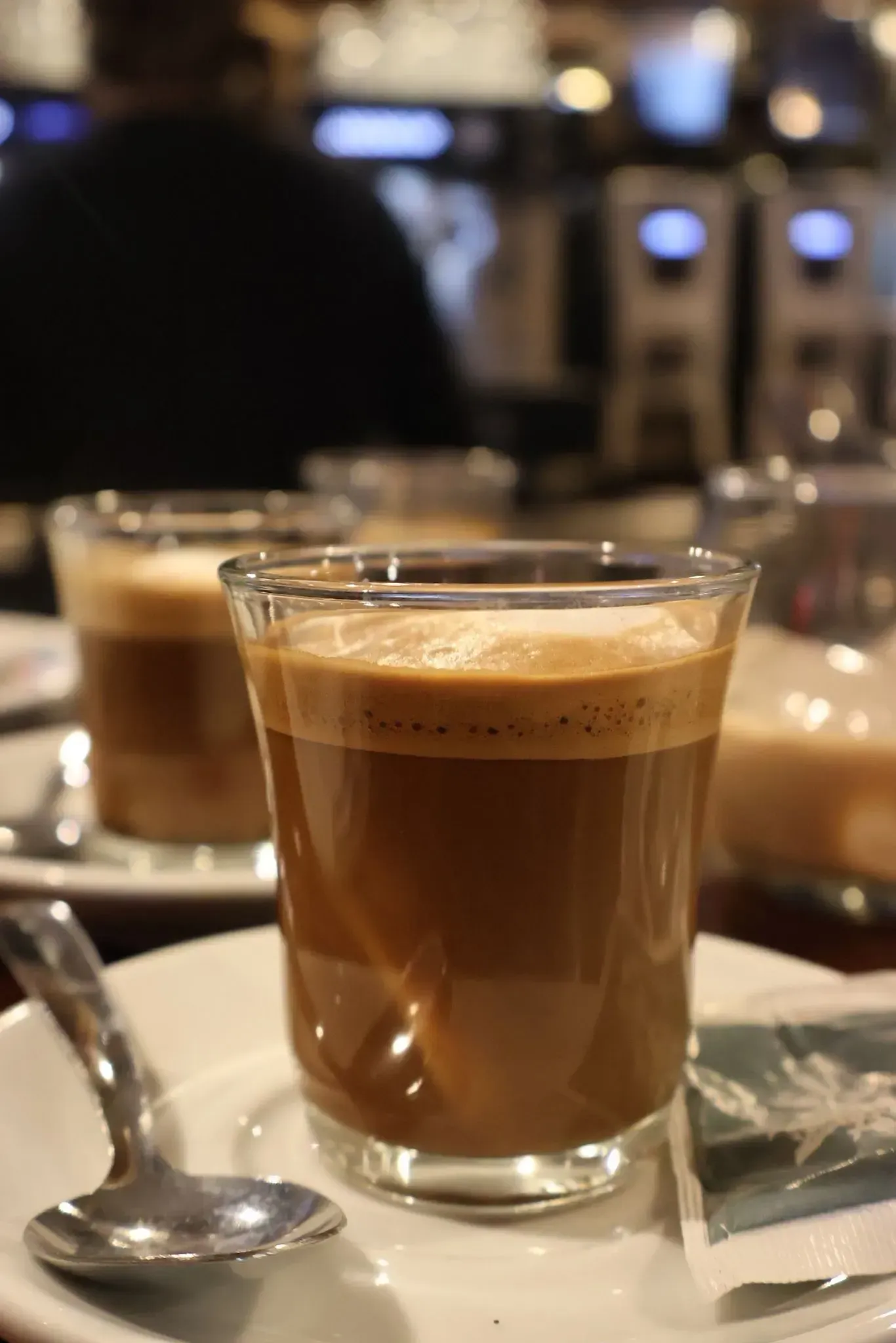 Primer plano de un vaso de espresso coronado con crema, servido en un platillo con una cuchara en una cafetería.